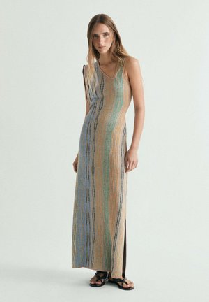 Mujer de pie con un vestido largo sin mangas, de rayas multicolor, con aberturas laterales y sandalias negras con tiras sobre un fondo liso.