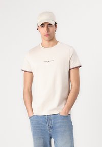 Beige bomull t-skjorte med rund hals, korte ermer og maroon-kantede kanter. Har en liten logo på brystet. Kombinert med lyse denim jeans.