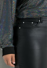 Zwarte faux leren broek met een voorkantzak en zilveren knoopaccenten, gecombineerd met een langarmtop bedekt met veelkleurige pailletten.