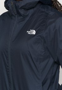 Tummansininen kevyt takki, jossa on huppu, etuvetoketju ja valkoinen logo rinnassa. Sileä, vettähylkivä kangas ja vartalonmyötäinen malli.