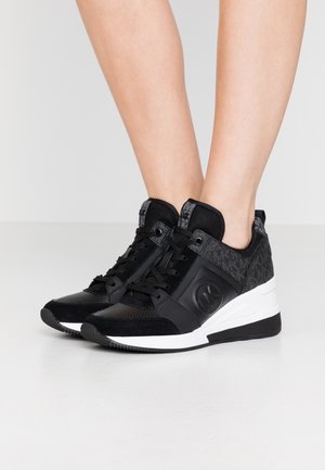 Person iført sorte og grå kilehælssneakers med hvide mellemsåler, stående på en lysegrå gulv mod en ensfarvet baggrund.