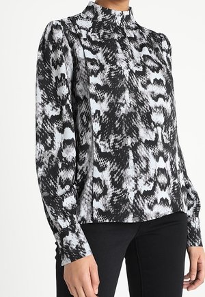 Schwarz-weiße Bluse mit Schlangenmuster, langärmlig und mit hohem Kragen, getragen mit schwarzer Hose, zeigt Oberkörper und Arme.