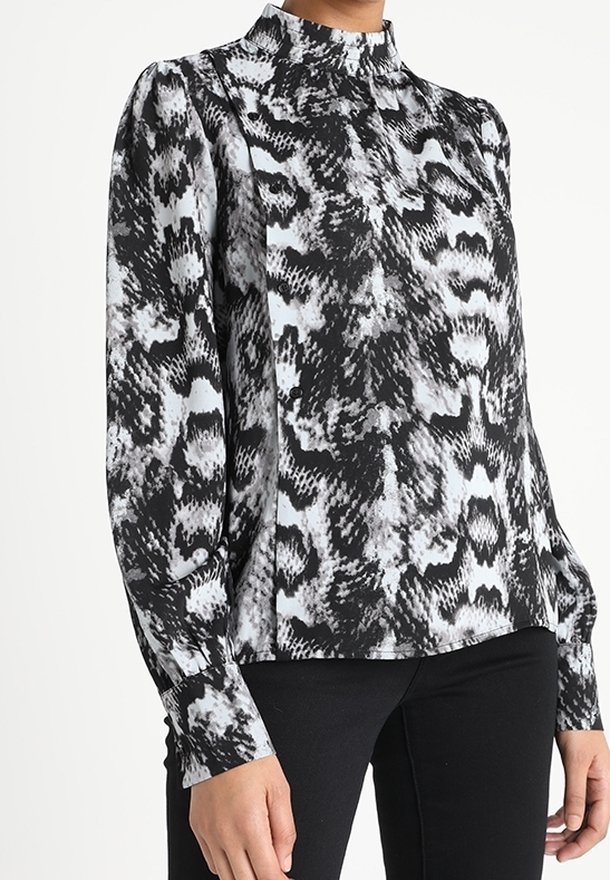 Blouse à manches longues à imprimé serpent noir et blanc avec col montant, portée avec un pantalon noir, montrant le haut du corps et les bras.