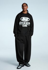 Čierna mikina s bielym grafickým textom, skombinovaná s čiernymi voľnými nohavicami a teniskami so striebornými akcentmi. Mäkkučká, štruktúrovaná tkanina.