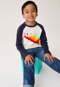 Bela in mornarsko modra majica z dolgimi rokavi z napisom "HAPPY" v živahnih barvah. Sedi na modrem bloku, oblečena v modre kavbojke. Gladek material, priloženski kroj.