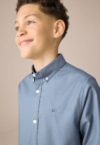 Camicia grigia con bottoni e colletto, con un piccolo logo di aragosta ricamato in blu. Tessuto di cotone, texture liscia e bottoni bianchi.