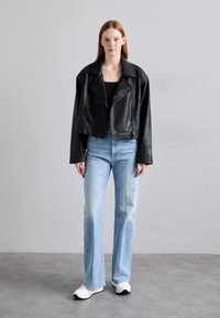 Schwarze Lederjacke mit weitem Revers, kombiniert mit hellblauen, ausgestellt geschnittenen Jeans und weißen Sportschuhen. Glatte Textur, minimale Hardware.