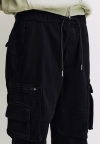 Pantaloni cargo negri cu talie reglabilă cu șnur, prevăzuți cu multiple buzunare și fermoare, realizați dintr-un material durabil. Textură distinctă, croială slim.