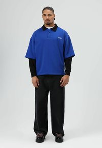 Blauw oversized poloshirt met een zwarte kraag, gecombineerd met zwarte jeans met wijde pijpen en zwarte schoenen met roze veters. Soepele stoftextuur.