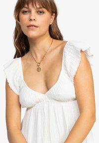 Bílá texturovaná šaty s řasenými rukávy a výstřihem do V. Zlatý řetízek s kulatým přívěskem doplňuje celý outfit.