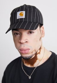 Svarte og hvite stripete baseballcaps med en gul logo-patch. Enkel svart t-skjorte og sølvsmykke synlig. Nærbilde portrettstil.