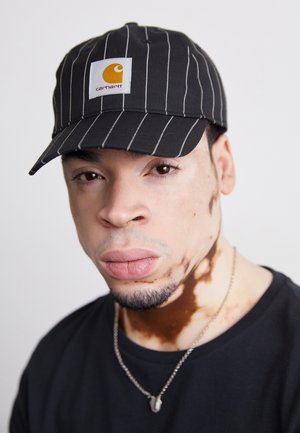 Black and white striped baseball cap with a yellow logo patch. Plain black t-shirt and silver necklace visible. Close-up portrait style.