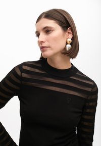 Čierny top s priesvitnými prúžkovanými rukávmi, rebrovaným golierom a malým vyšitým logom na hrudi. Má hladkú textúru a priliehavý strih.