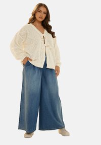 Krämfärgad stickad cardigan med diamantmönster, knuten fram, tillsammans med löst sittande blå denim byxor i bredbent modell och beige sneakers.