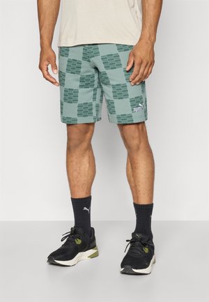 Mand iført grønne Puma-shorts med gentaget logo mønster, sorte Puma-strømper og sorte og hvide sportssko, der står mod en hvid baggrund.