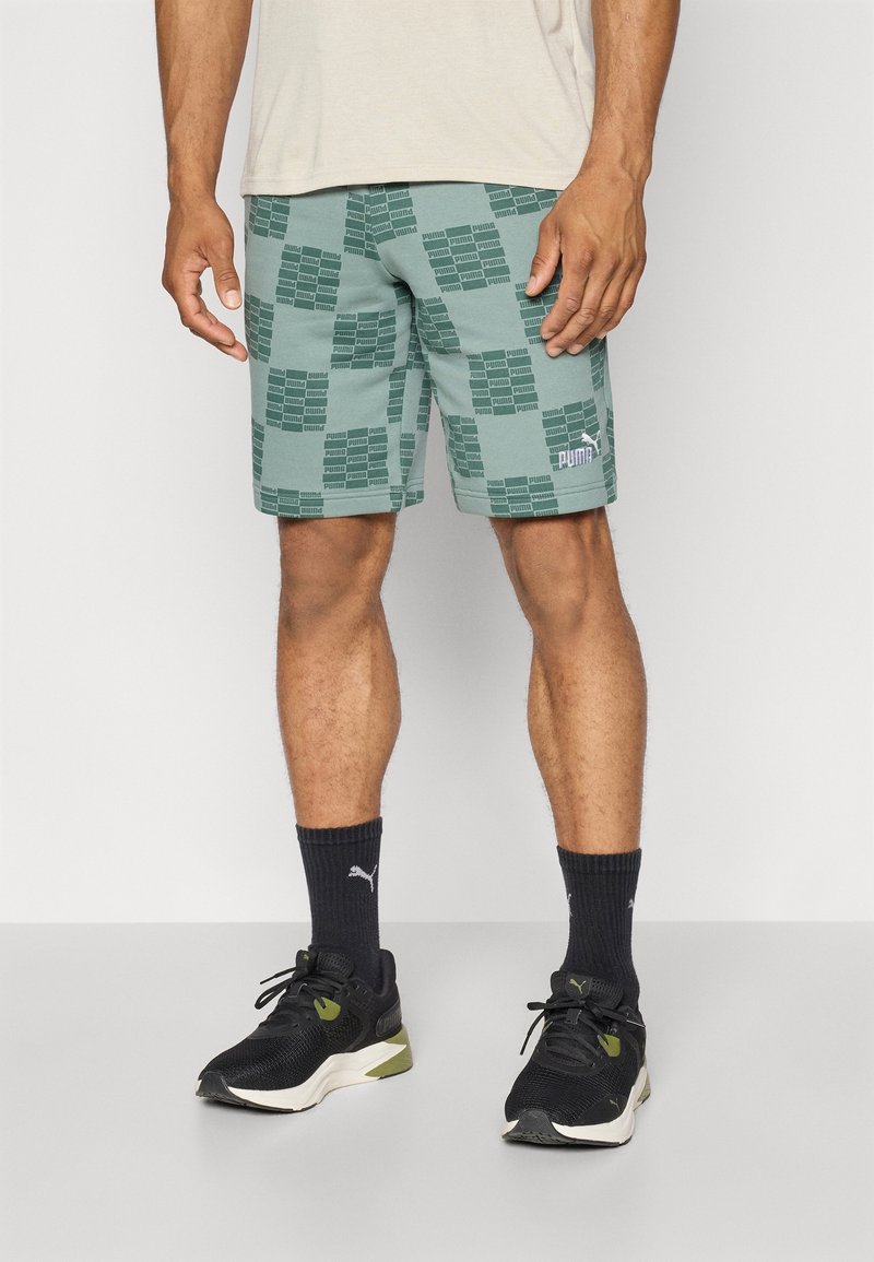 Homme portant un short vert Puma avec un motif de logo répété, des chaussettes Puma noires et des chaussures de sport noires et blanches, debout sur fond blanc.