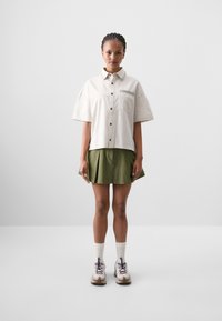 Lys beige kortermet skjorte med dekorativ krage, brystlomme og brune knapper, kombinert med et grønt folderett skjørt og joggesko.