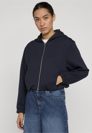 Kvinde med mørkt hår sat op i en hestehale iført en marineblå hættetrøje med lynlås og blå jeans, stående mod en ensartet lys baggrund.