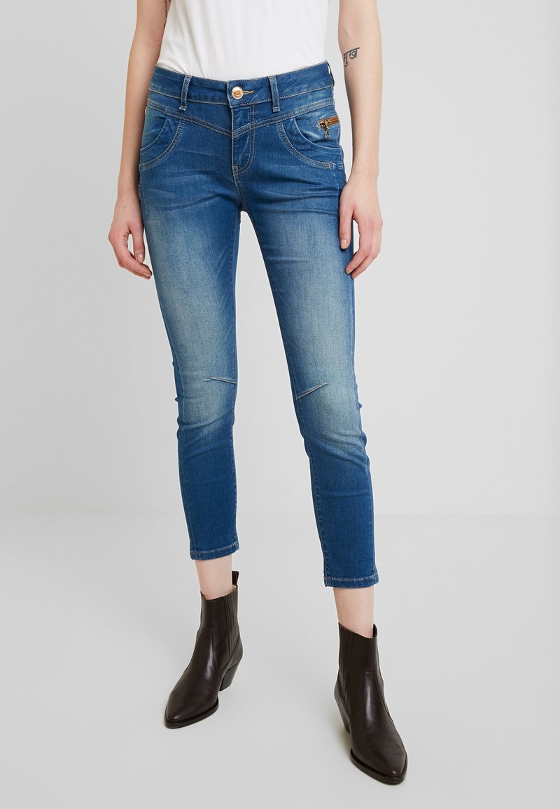 Jean en denim bleu avec une coupe slim, taille mi-haute, cinq poches, détails de décoloration légère et une poche zippée à l'avant. Accompagné de bottines marron.
