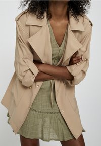 Beige trench coat with rolled sleeves, gold accents, and a loose fit, layered over a light green dress with a textured, tiered skirt.