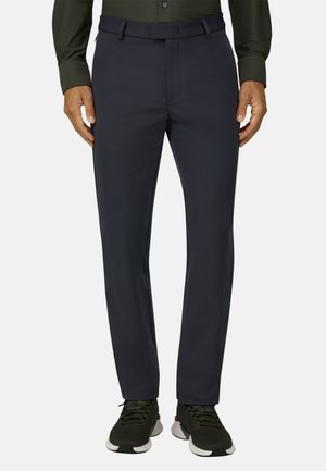 Homme portant un pantalon noir slim, des baskets noires à semelles blanches et une chemise à manches longues vert foncé, debout devant un fond uni.
