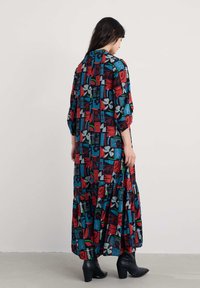 Długa, wzorzysta sukienka z wyrazistymi czerwonymi, niebieskimi i szarymi geometrycznymi kształtami. Miękka tkanina, rękawy o długości trzech czwartych oraz warstwowy krój spódnicy.