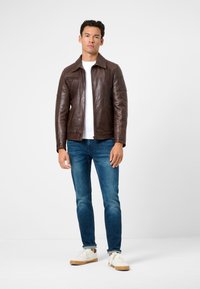 Bruine leren jas met een gespreid kraag, een ritszak op de torso en een gladde textuur, gedragen over een wit shirt en blauwe spijkerbroek.