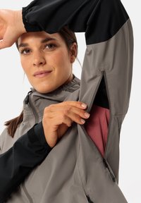 Giacca leggera grigia con dettagli neri, dotata di una tasca laterale con zip e fodera in rete rosa, e polsini elastici per una vestibilità sicura.