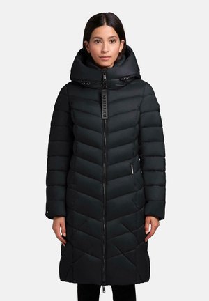 Vrouw in een lange zwarte gewatteerde winterjas met hoge gewatteerde kraag en ritssluiting aan de voorkant, staande tegen een effen achtergrond.
