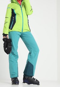 Světlá zelená a černá lyžařská bunda s modrými akcenty, doplněná světlomodrými lyžařskými kalhotami s tmavě modrým panelem, nošena s černými lyžařskými botami.