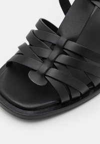 Sandale noire avec des lanières en cuir tressé, semelle plate et texture lisse, conçue pour un port ouvert avec des accents minimes.