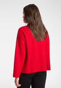Roter, übergroßer Pullover aus weichem Stoff. Verfügt über eine lockere Passform, lange Ärmel und einen abgerundeten Saum. Getragen mit schwarzer Hose.
