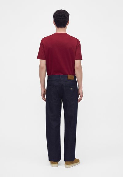 Hombre de pie de espaldas, vestido con una camiseta roja, vaqueros azul oscuro con bolsillos traseros y zapatos casuales color beige sobre un fondo blanco liso.
