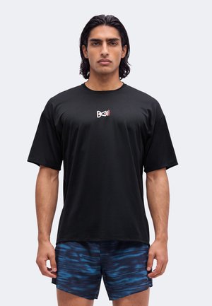 Homme aux cheveux noirs mi-longs porte un t-shirt noir avec un petit logo blanc et rouge et un short bleu à motifs, debout face à l'avant.