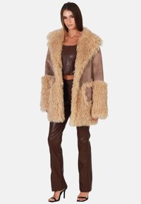 Bruine schapenvachtjas met bontafwerking en grote zakken, gedragen over een bruin crop-top en faux leren broeken, met open teen zwarte hakken.