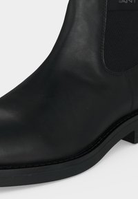 Botte Chelsea en cuir noir avec panneau latéral élastique et talon bas empilé sur fond blanc.