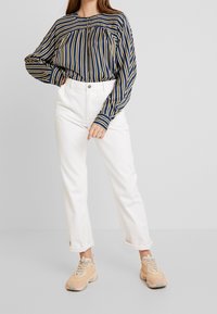 Chemise à manches longues rayée bleu marine et jaune ; pantalon blanc taille haute ; baskets beige avec semelle grise ; coupe décontractée, poignets retroussés.