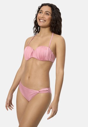 Mujer sonriente con cabello rizado, usando un bikini rosa con parte superior fruncida y detalle recortado en la parte inferior, posando frente a un fondo liso.