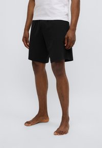 Shorts negros hechos de mezcla de algodón, con una cintura elástica, corte recto y sin patrones o detalles visibles, usados descalzo.