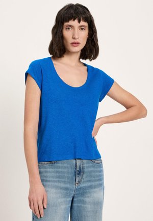 Woman with shoulder-length dark hair wearing a bright blue short-sleeve scoop neck top and light blue jeans, standing with hand on hip.