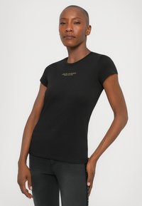 Vrouw met kort haar in een aansluitend zwart Armani Exchange t-shirt en donkere jeans, staande tegen een effen lichte achtergrond.