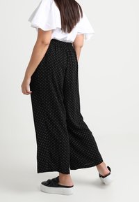 Pantalon noir à jambes larges avec des pois blancs, doté d'une taille élastique. Associé à un haut blanc à volants et des chaussures noires sans lacets.