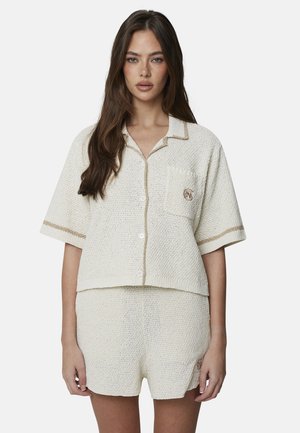 Vrouw met lang bruin haar, gekleed in een roomkleurig, gestructureerd kortemouw-blouseje met knopen en bijpassende shorts met subtiele beige randdetails.