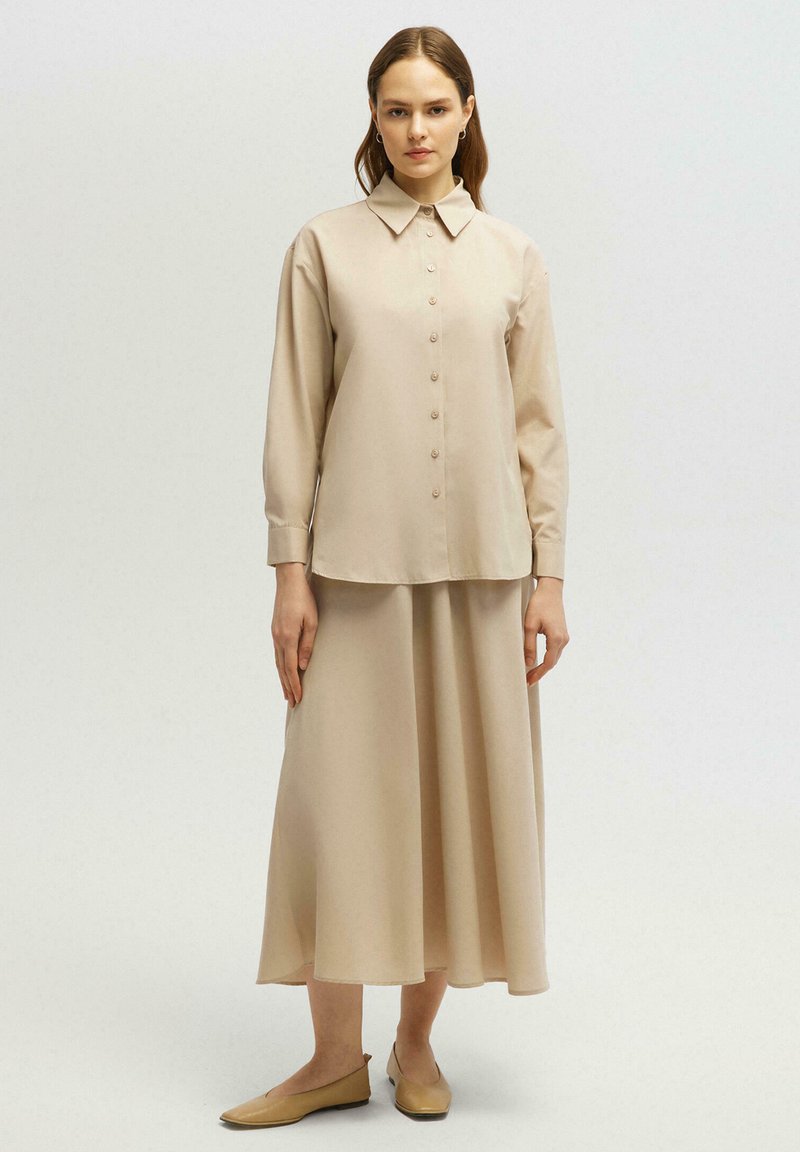 Chemise beige à manches longues, associée à une jupe évasée assortie. Tissu doux avec une texture lisse. Chaussures plates beiges discrètes.