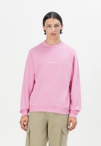 Sweat-shirt rose clair avec des manches longues, col rond et poignets côtelés. Présente un logo brodé sur la poitrine. Porté avec un pantalon cargo beige.