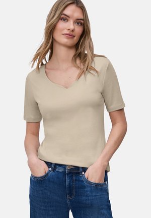 Kobieta o blond włosach do ramion, ubrana w beżową bluzkę z krótkim rękawem i niebieskie dżinsy, ręce w kieszeniach, zwrócona przodem.