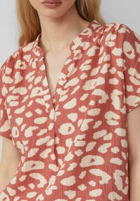 Kurzärmlige, kragenlose Bluse in Koralle mit einem cremefarbenen, abstrakten Leopardenmuster. Der Stoff hat ein strukturiertes, leichtes Aussehen.