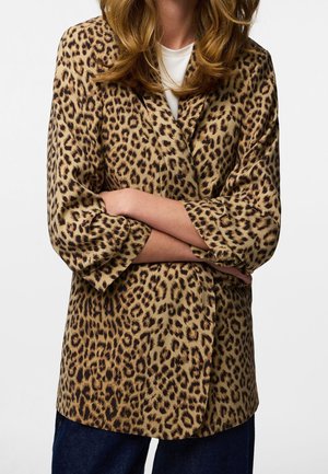 Frau trägt Leopardenmuster-Blazer mit hochgekrempelten Ärmeln und verschränkten Armen, kombiniert mit dunkelblauen Jeans und einem weißen Top darunter.