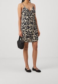 Robe portefeuille noire et beige avec un motif géométrique de feuilles, fines bretelles réglables et tissu léger. Associée à un sac noir matelassé et des ballerines noires.