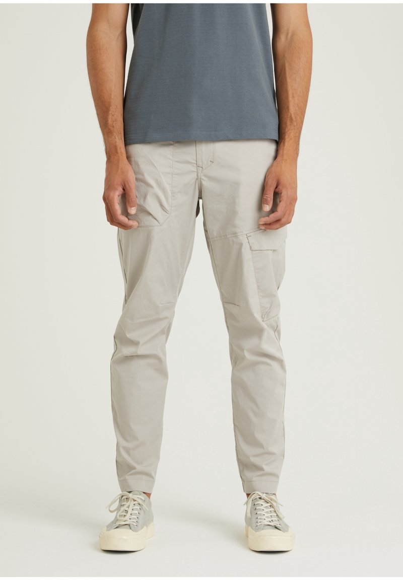 Homme portant un pantalon cargo beige clair et des baskets grises, debout devant un fond uni, mains détendues le long du corps.